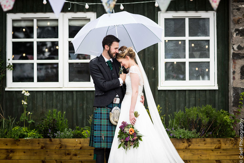 Bride and Groom embrace under white umberella outside Comrie Croft Wedding Venue