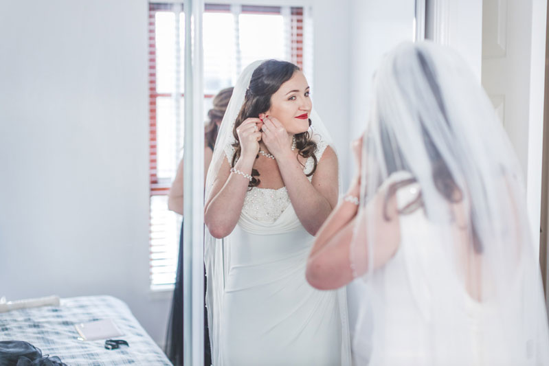 Philippa in her wedding dress and veil placing earrings